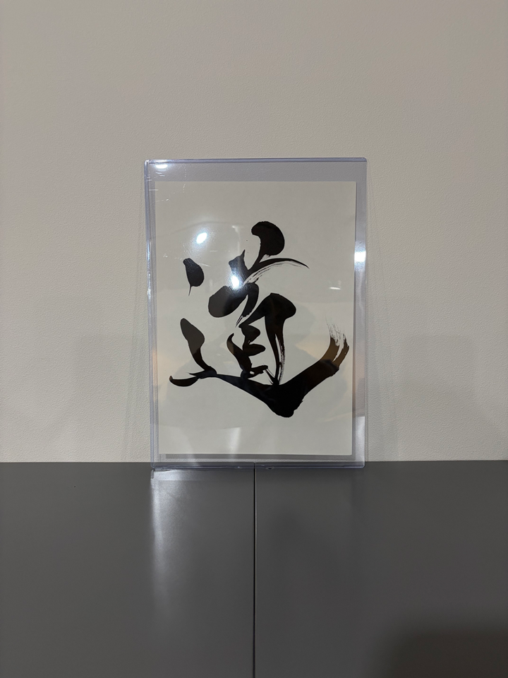 Do (道), The Way, in black brush calligraphy on white paper, displayed in a clear frame on a dark tabletop.